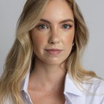 A confident blonde woman posed against a neutral background wearing a white shirt.
